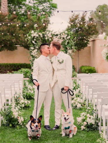 Grooms kiss with dogs at outdoor wedding ceremony.