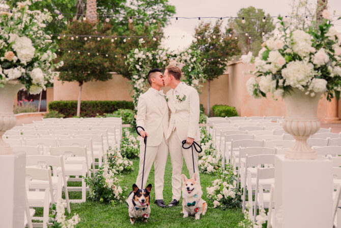Grooms kiss with dogs at outdoor wedding ceremony.