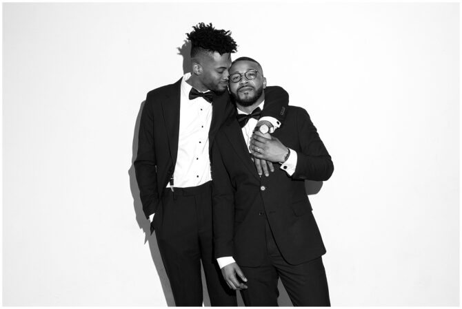 Two men in tuxedos embrace, smiling warmly.