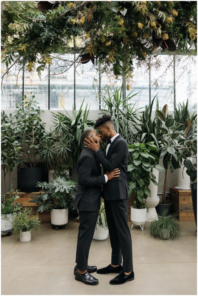 Couple in suits kissing in greenhouse setting.
