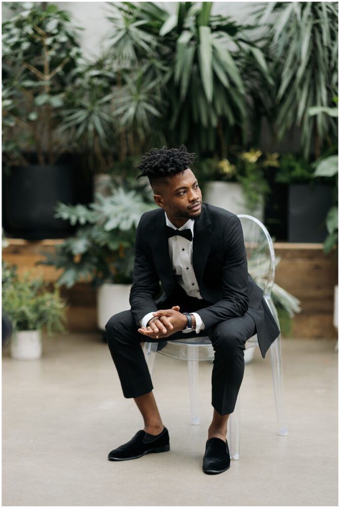 Man in tuxedo sitting, surrounded by plants.