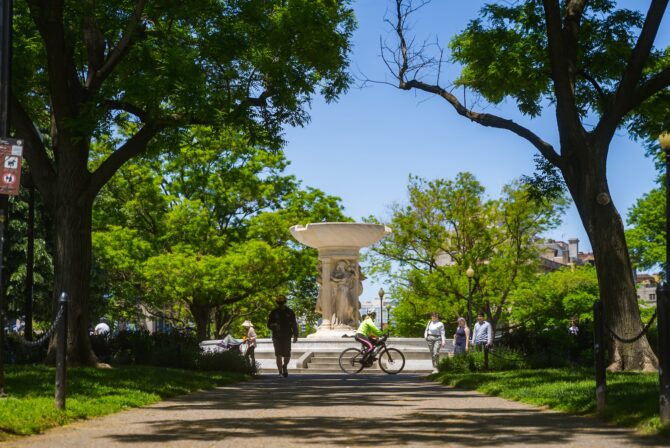 Park scene with fountain and trees, cyclists and pedestrians.